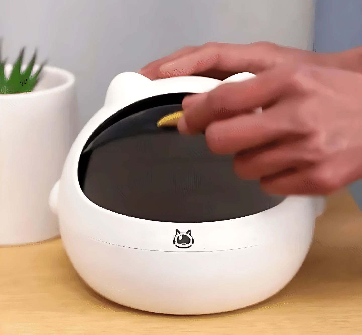 Mini basurera blanca con tapa estilo gato, manos depositando objeto en interior.