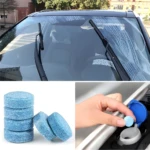Pastillas efervescentes azules para limpia parabrisas automotriz, apiladas y siendo introducidas en depósito de auto.