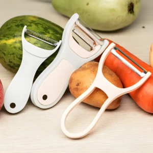 Set x2 peladores de frutas y verduras 360°, uno blanco y otro rosado, con papas, zanahorias y calabaza de fondo.