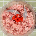 Picadora manual de alimentos con carne molida rosa picada y cuchillas grises y rojas dentro de un recipiente de vidrio.