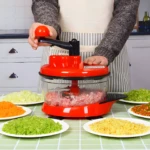 Picadora de alimento manual roja en uso, rodeada de platos con vegetales picados y carne molida.