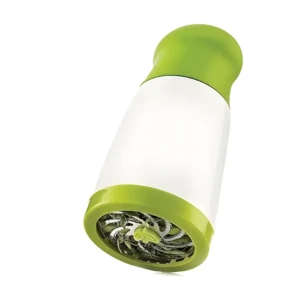 Picadora manual de hierbas verde y blanca en ángulo inferior, con cuchillas expuestas y trozos de hierbas verdes.