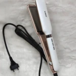 Plancha alisadora VGR blanca con bordes dorados y cable negro, apoyada sobre una superficie de peluche blanca, mostrando placas lisas y control térmico.