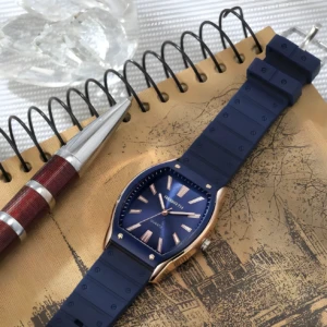 Reloj Rebirth de cuarzo con caja dorada, esfera azul y correa de silicona azul, posado sobre una libreta de notas con una pluma elegante.