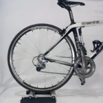 Bicicleta de ruta blanca y negra en rodillo de entrenamiento, vista lateral trasera.
