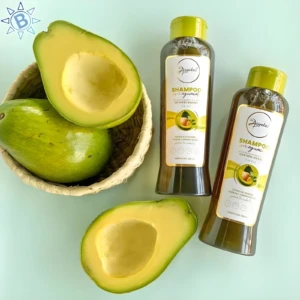 Dos envases de Shampoo Anyeluz con aguacate de 500 ml junto a una canasta con aguacates frescos enteros y partidos sobre una superficie verde clara.