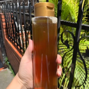 Botella con shampoo artesanal dorado, ideal para realzar reflejos en cabello claro, sostenida por mano con fondo exterior.