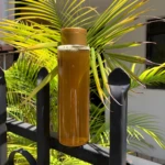 Botella de shampoo artesanal de manzanilla con tapa dorada, líquido ámbar espumoso, en reja negra, hojas verdes y amarillas al fondo.