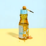 Botella de shampoo Milagros Kids color ámbar con diseño de animales, sobre superficie amarilla y fondo azul claro, con dosificador y gota de miel.