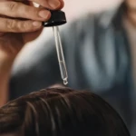 Mano aplicando suero capilar anti-piojos transparente directamente sobre el cuero cabelludo mediante un gotero de vidrio con tapa negra.