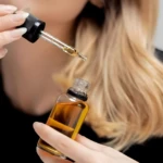 Persona sosteniendo un frasco de suero capilar color ámbar y un gotero lleno con el mismo tratamiento líquido, diseñado para el cuidado y tratamiento capilar.