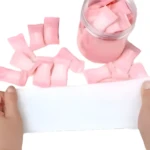 Toallas compactas de limpieza facial rosa, apiladas y en frasco de vidrio, sujetas con manos sobre superficie blanca.