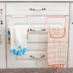 Set de 2 toalleros colgantes blancos para cocina, uno con guantes y otro con paño a cuadros rosa y naranja.