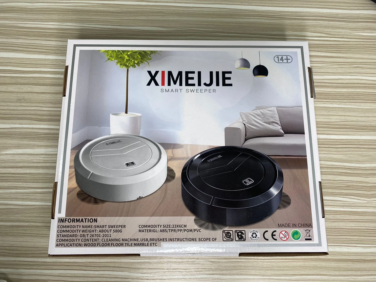 Robot aspirador recargable XIMEIJIE en caja, mostrando dos unidades: una blanca y otra negra, sobre suelo de madera con sofá y planta al fondo.