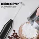 Batidora portátil negra para café con varilla metálica espumando leche en vaso, junto a granos de café.