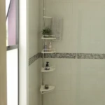 Estante esquinero telescópico blanco para ducha con cuatro repisas triangulares, barra metálica ajustable, toallero y accesorios organizados sobre azulejos claros