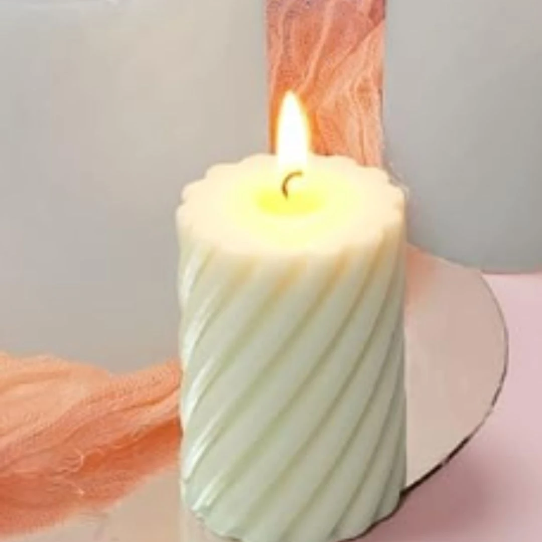 Vela artesanal blanca con diseño espiral en espiral, iluminada con llama cálida, sobre fondo rosa y tela coral.