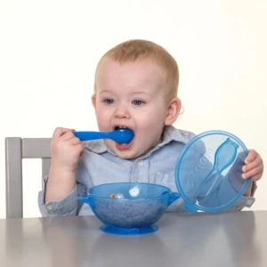 Bebé azul comiendo cereal con cuchara de un plato antiderrame con ventosa y tapa.