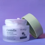 Crema facial Centella Asiática BIOAQUA en envase blanco translúcido con tapa verde claro abierta, fondo morado.