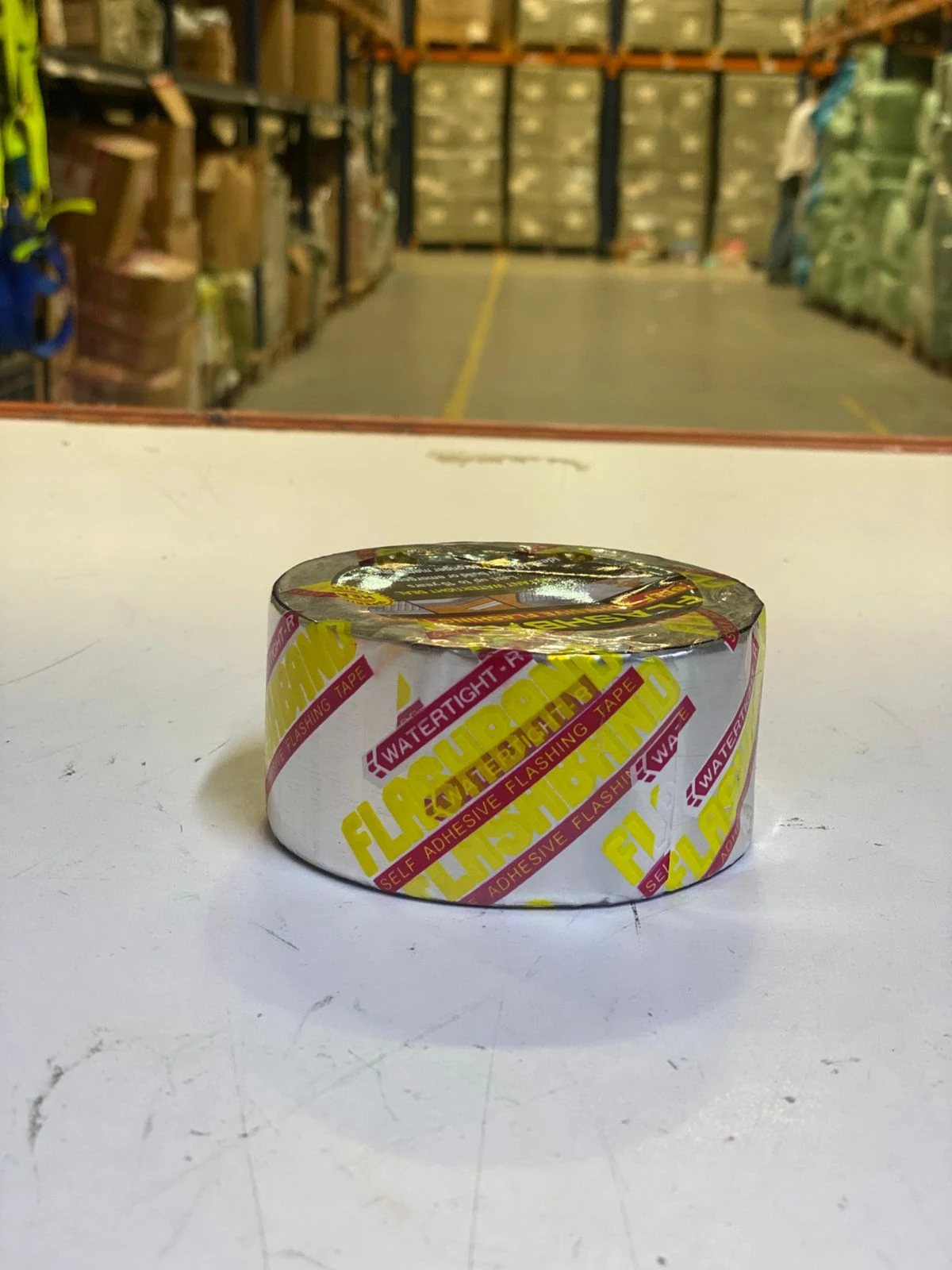 Cinta de aluminio autoadhesiva impermeable, rollo con diseño en amarillo y rojo sobre fondo blanco, sobre superficie blanca en almacén.