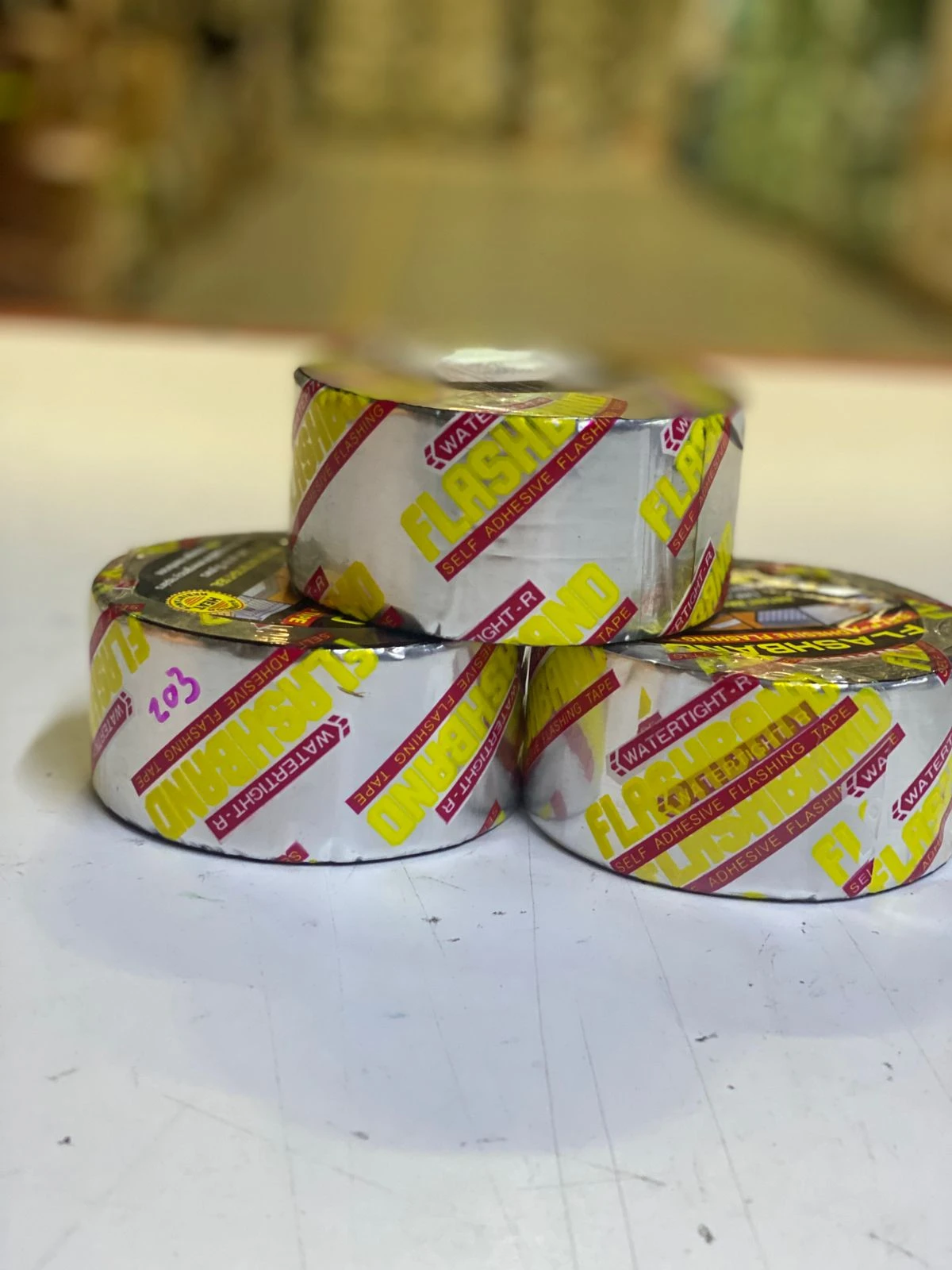 Tres rollos de cinta de aluminio autoadhesiva, color plateado con etiquetas amarillas y rojas, apilados sobre superficie blanca.