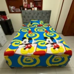 Sábana infantil con estampado de Mickey Mouse en azul, amarillo y rojo, sobre cama con cabecero gris tapizado.