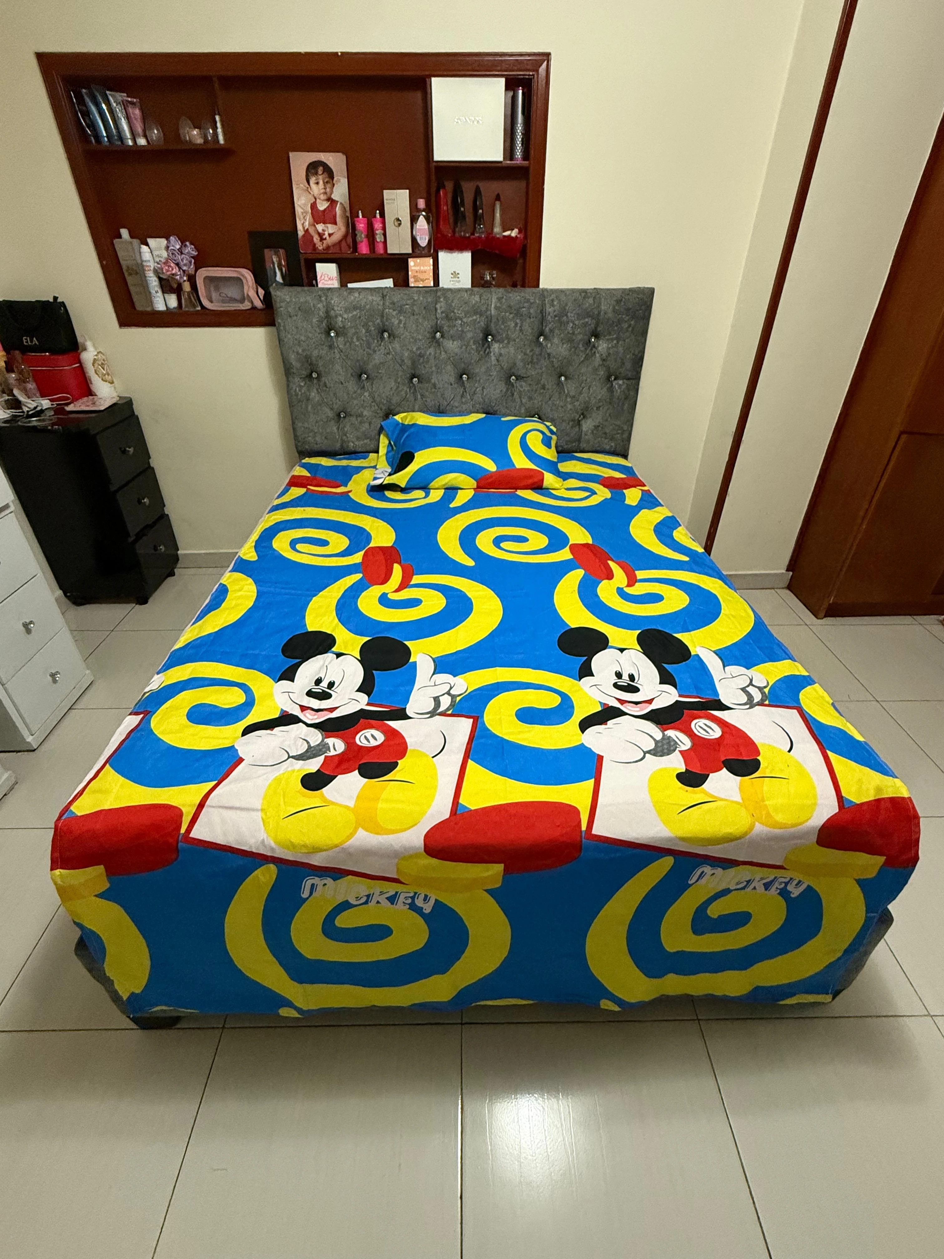 Sábana infantil con estampado de Mickey Mouse en azul, amarillo y rojo, sobre cama con cabecero gris tapizado.