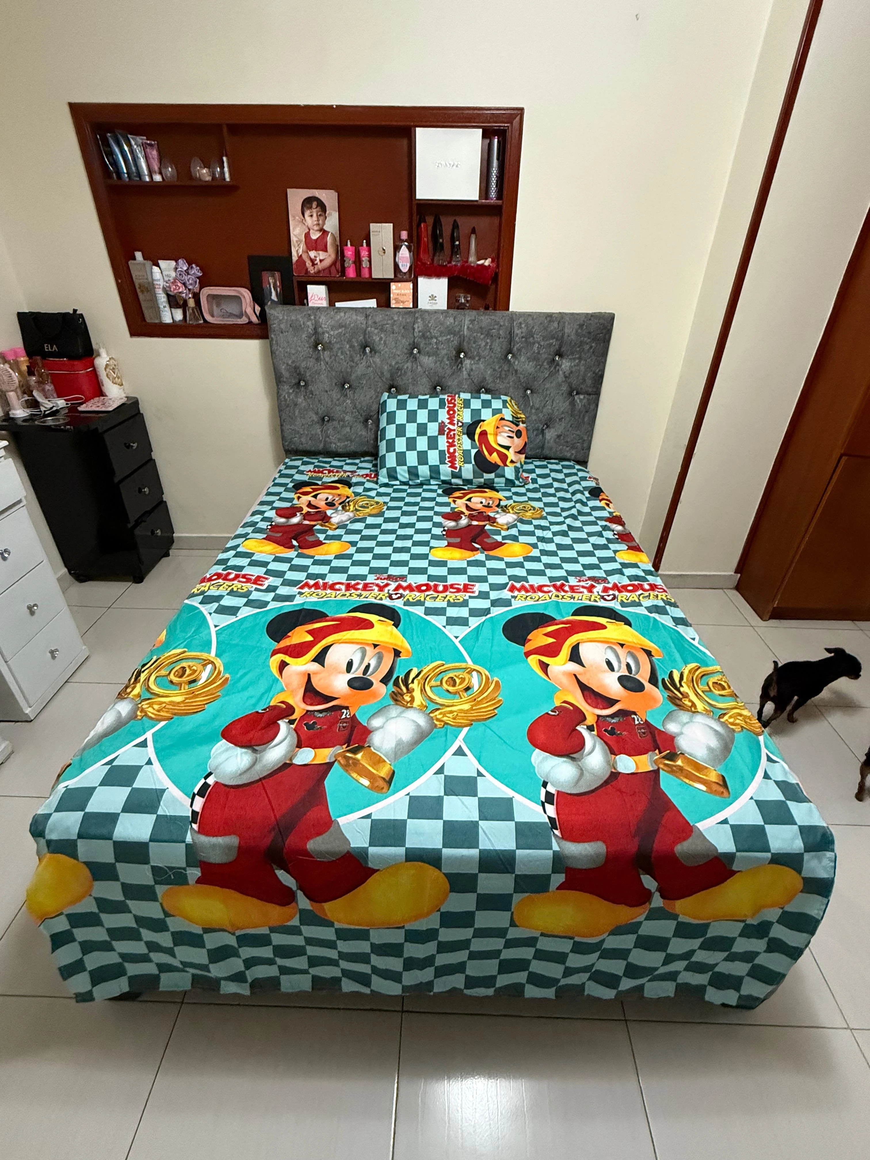 Sábana infantil con estampado de Mickey Mouse en tonos turquesa y rojo, sobre cama con cabecero gris tapizado.