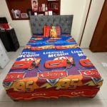 Sábana infantil de tema Cars con estampado de Rayo McQueen, colchón gris tapizado y estantería de madera al fondo.