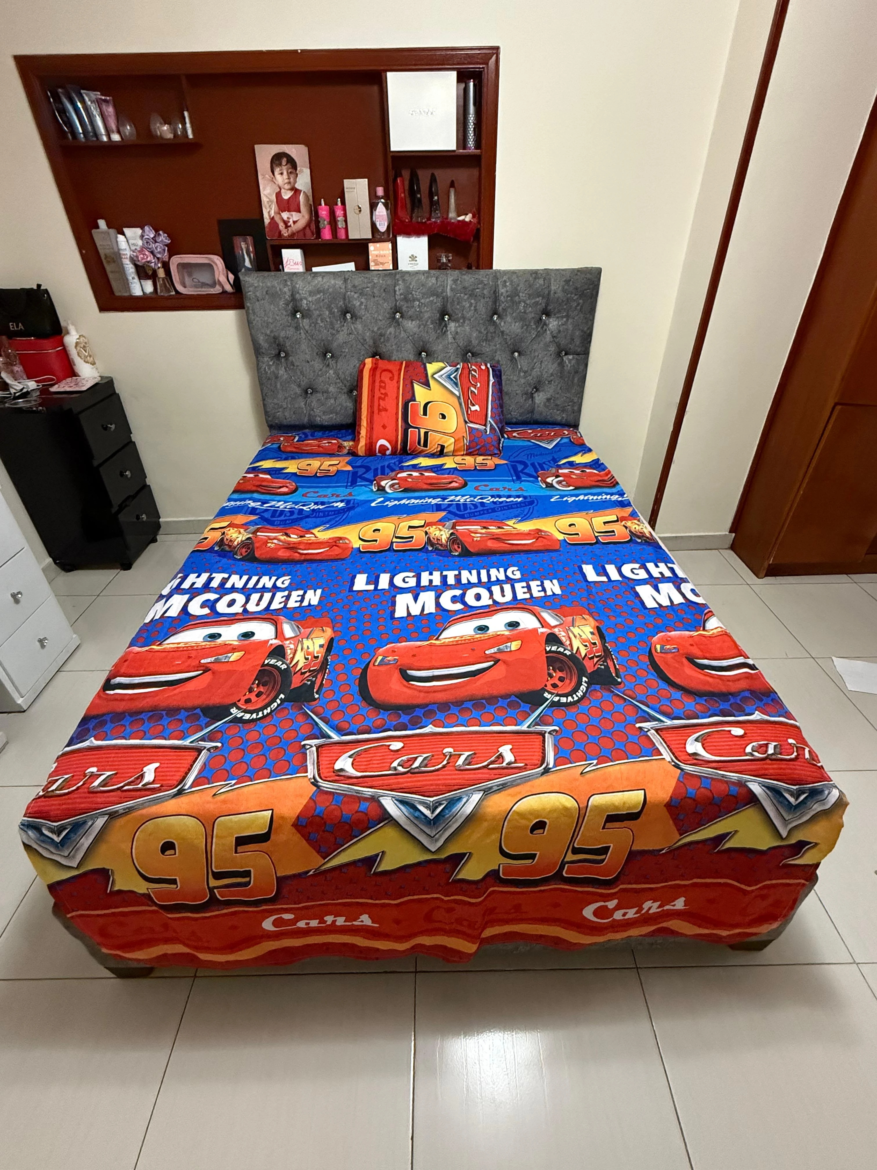 Sábana infantil de tema Cars con estampado de Rayo McQueen, colchón gris tapizado y estantería de madera al fondo.