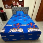 Sábana infantil azul con estampado de Spider-Man, sobre cama con cabecero tapizado gris en habitación.