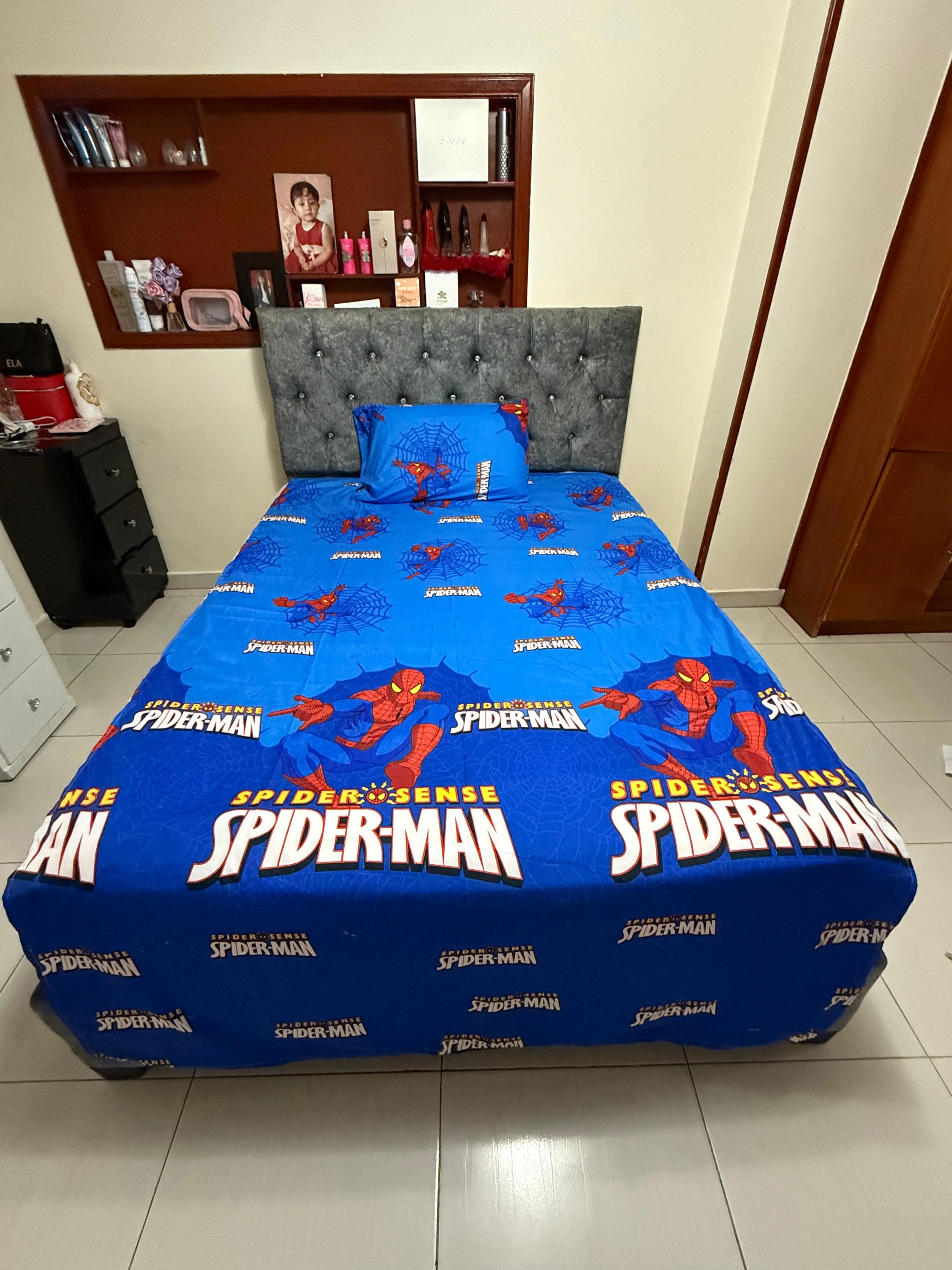Sábana infantil azul con estampado de Spider-Man, sobre cama con cabecero tapizado gris en habitación.