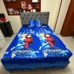 Sábana infantil Frozen de algodón suave, con estampado de Elsa y Anna en azul vibrante, sobre cama con cabecero tapizado gris.