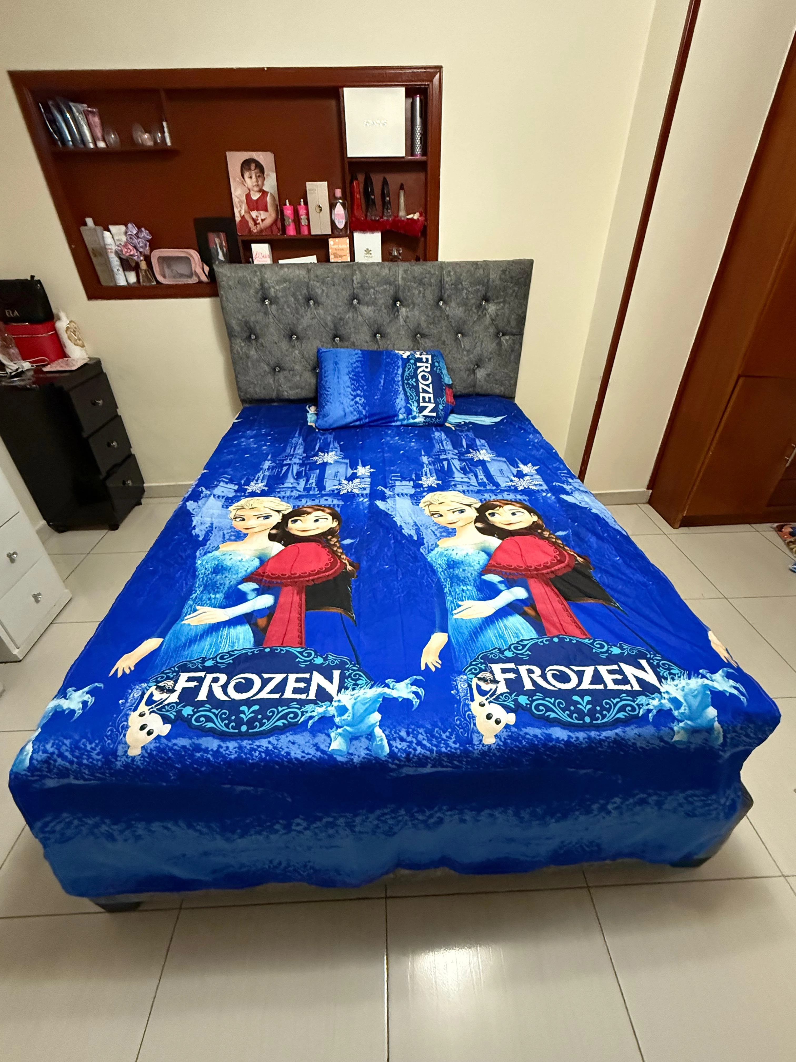 Sábana infantil Frozen de algodón suave, con estampado de Elsa y Anna en azul vibrante, sobre cama con cabecero tapizado gris.