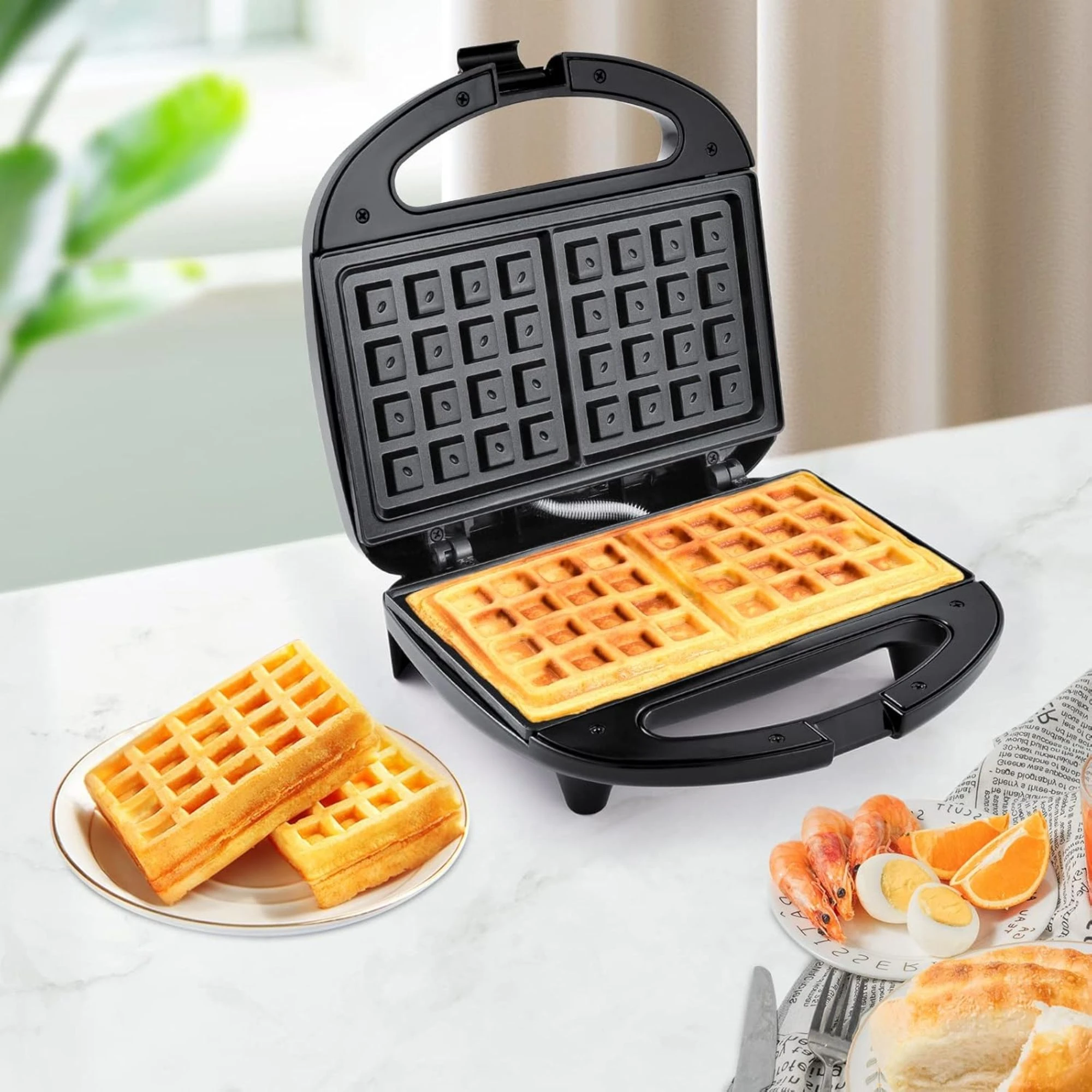 Sandwichera waflera negra abierta, mostrando moldes internos y waffles dorados dentro; acompañada de waffles en plato y alimentos frescos sobre mesa blanca.