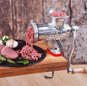 Molino de carne manual de aluminio Victoria, en uso sobre tabla de madera, extruyendo carne picada sobre plato negro con ingredientes frescos.