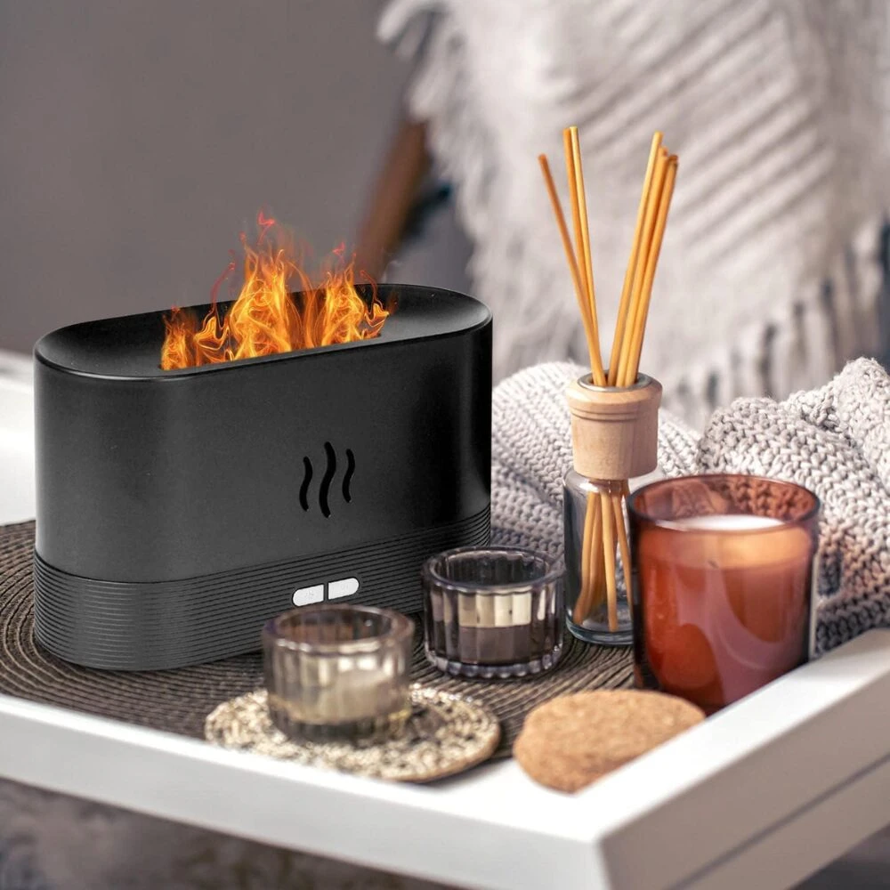 Difusor ultrasónico negro con llama realista y varillas aromáticas, sobre bandeja con velas y cojín, ideal para ambiente relajante.