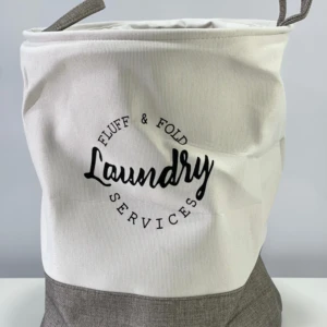 Cesto de tela para ropa sucia, blanco con base gris, diseño minimalista con texto "Laundry Services".