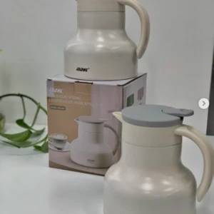 Jarra térmica MAHL en tono crema con tapa gris, modelo Mountain Spring. Se muestra una unidad sobre su caja de cartón y otra al frente, destacando su diseño ergonómico y asa curva sobre superficie blanca.