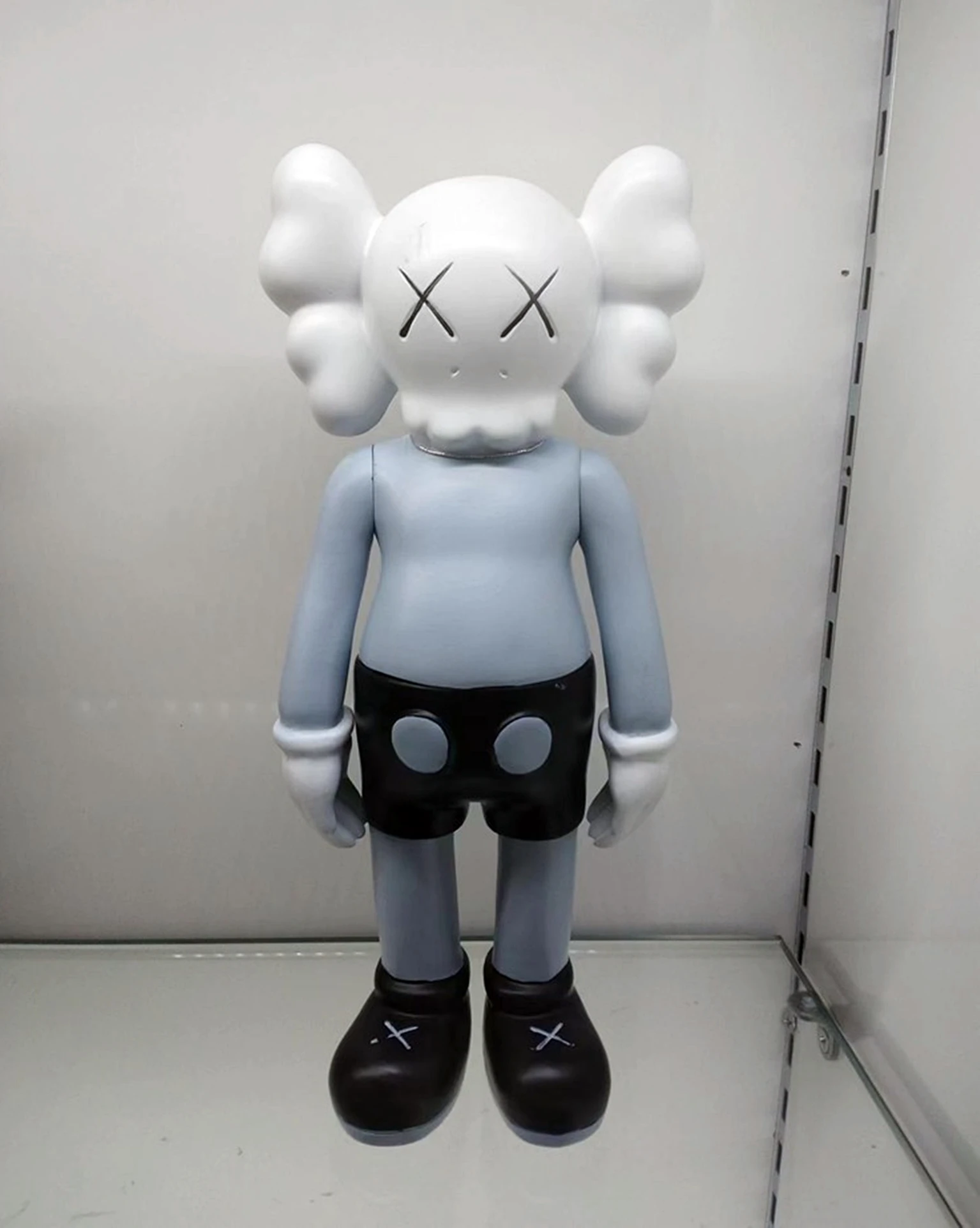 Figura decorativa de resina estilo KAWS de 18 cm, con cabeza blanca, ojos en forma de X, cuerpo gris claro, shorts negros con círculos grises y zapatos negros con X.