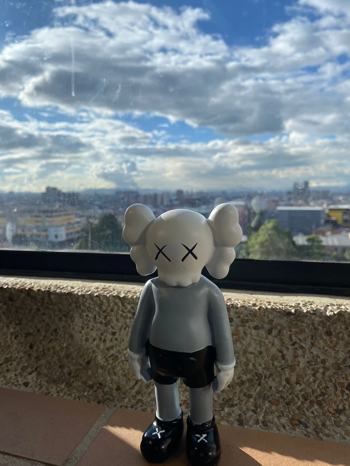 Figura decorativa de resina estilo KAWS, color gris con ojos en X, 18 cm, sobre alféizar con vista urbana.
