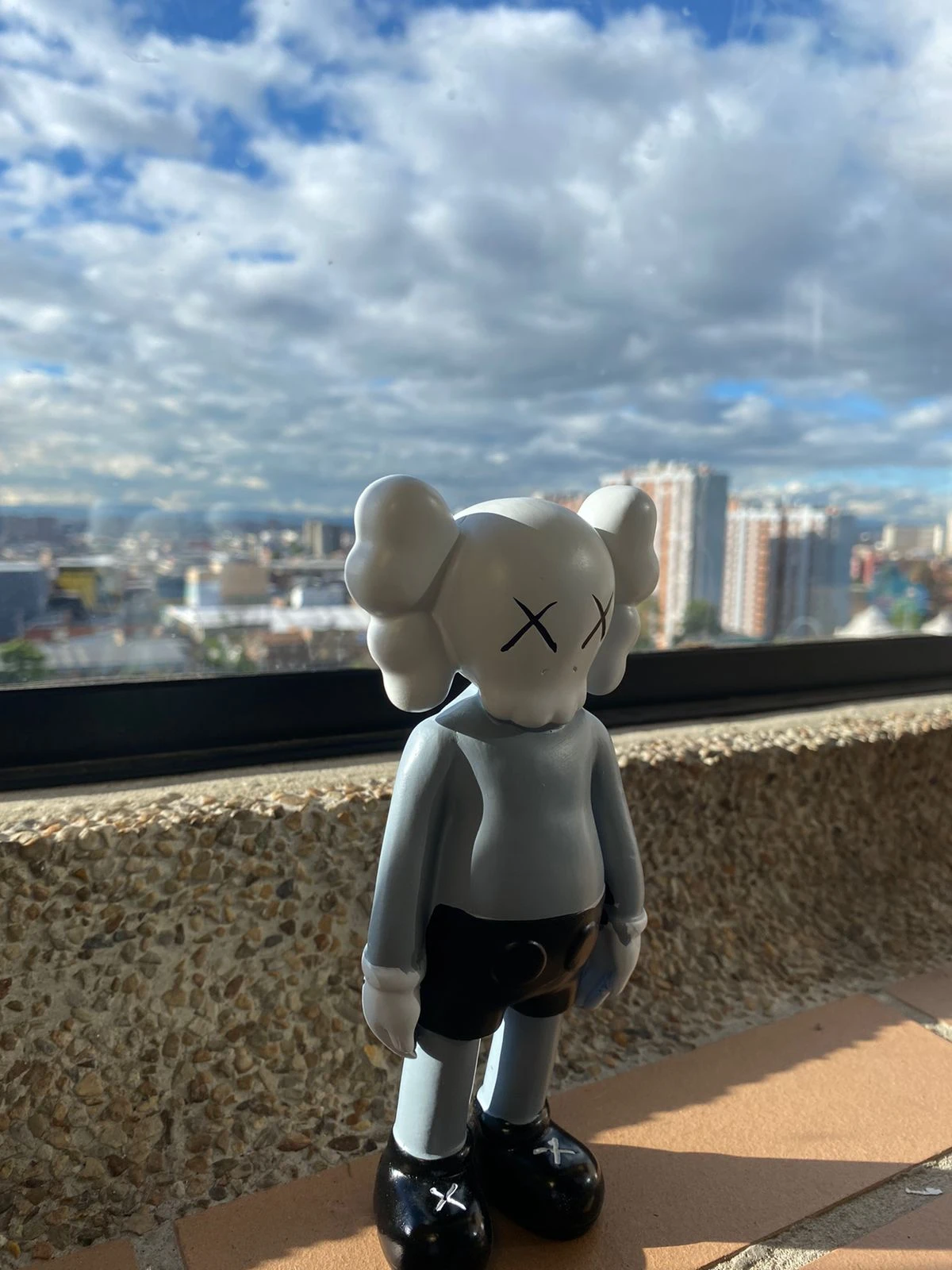 Figura decorativa de resina estilo KAWS de 18 cm, con acabado gris y detalles en negro, colocada sobre una superficie de baldosa frente a una ventana con vista urbana bajo cielo parcialmente nublado.