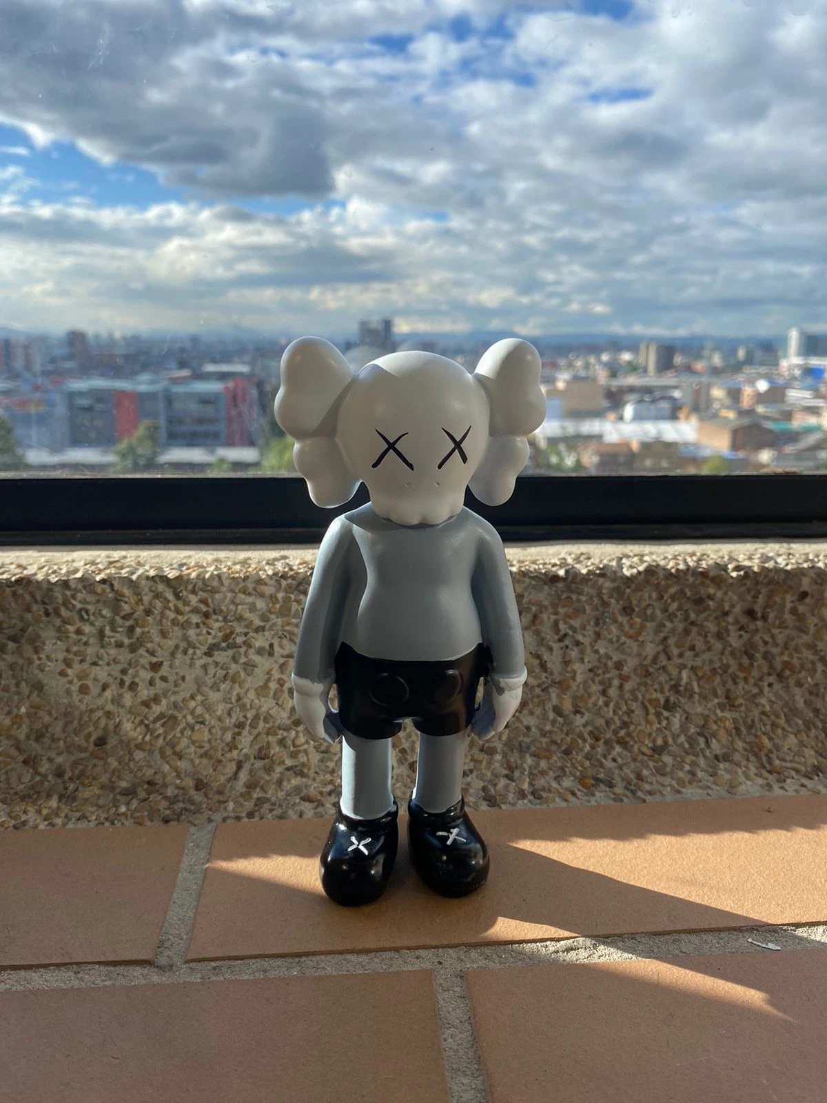 Figura decorativa de resina estilo KAWS, color gris claro con detalles negros, ojos en forma de X, sobre fondo urbano.