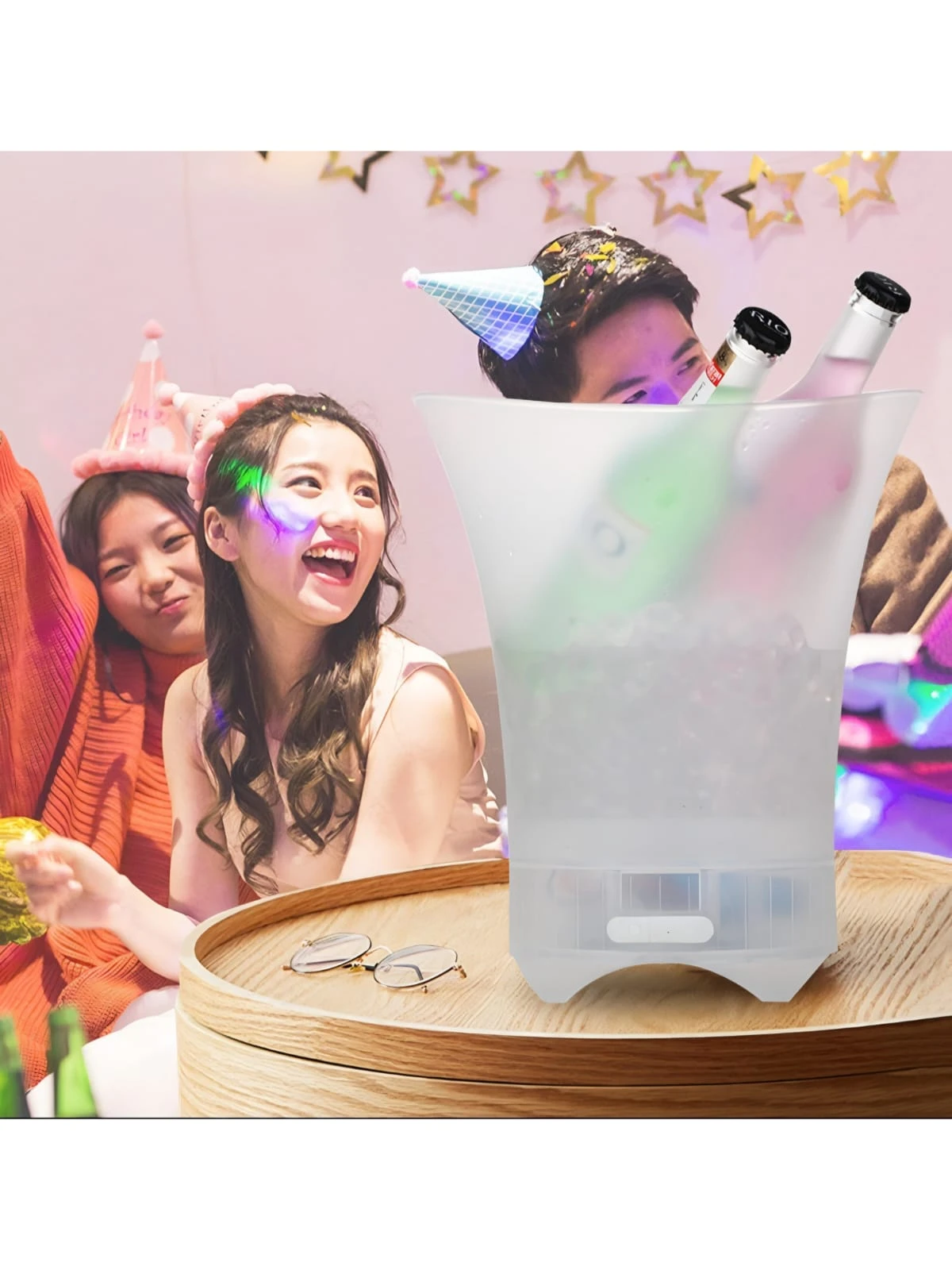 Cubeta hielera blanca con luz LED y altavoz Bluetooth, sobre mesa de madera, con botellas de bebida y personas celebrando al fondo.