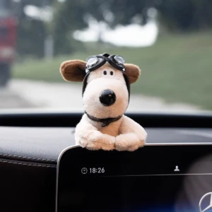 Peluche de perro de pelaje beige con gafas de piloto y corbata negra, colocado sobre el salpicadero de un coche. Vista frontal desde el interior del vehículo.