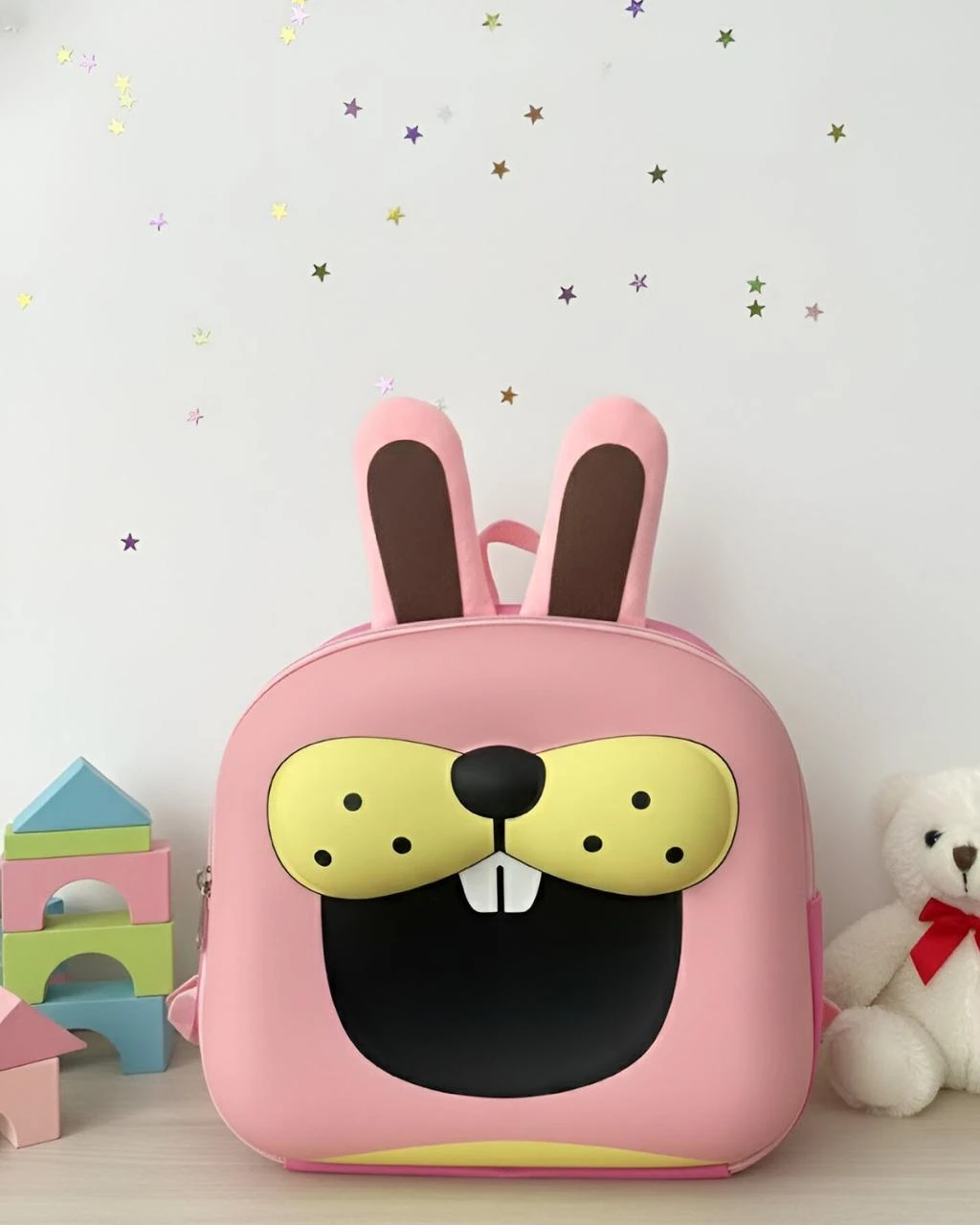 Mochila rosa con diseño de conejo, orejas erguidas y cara amarilla con dientes blancos, sobre fondo claro con juguetes infantiles.