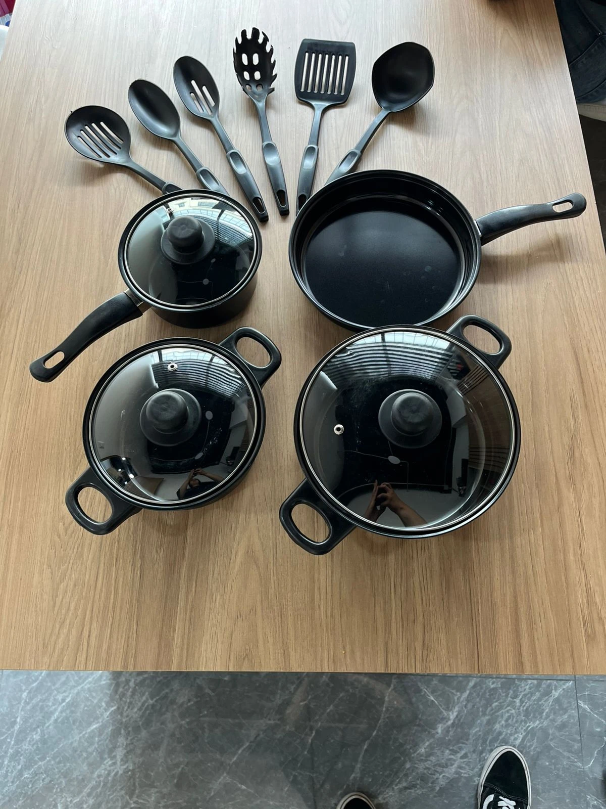 Set de cocina negro con ollas, sartén y utensilios sobre mesa de madera.