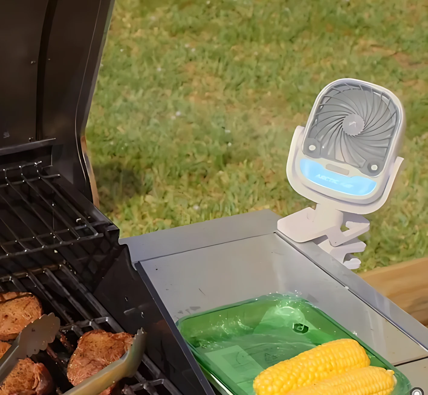 Difusor de aire portátil Arctic Air Grip Go con pinza, blanco, con luz azul, sobre parrilla con carne y maíz.