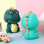 Alcancía de dinosaurio verde y turquesa, con detalles naranjas, ideal para niños.