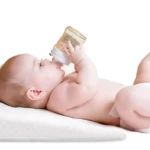 Bebé en pañal tomando tetero sobre almohada antireflujo blanca para colecho, previene regurgitación.